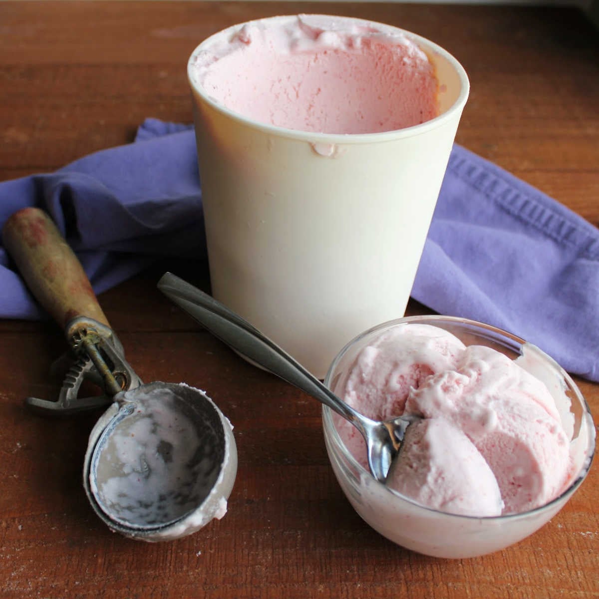 Homemade Strawberry Ice Cream Without Eggs 5 Antique ice cream scoop next to a freezer container filled with homemade strawberry ice cream and a small glass bowl filled with two scoops of creamy ice cream with a bite on a spoon, ready to eat.