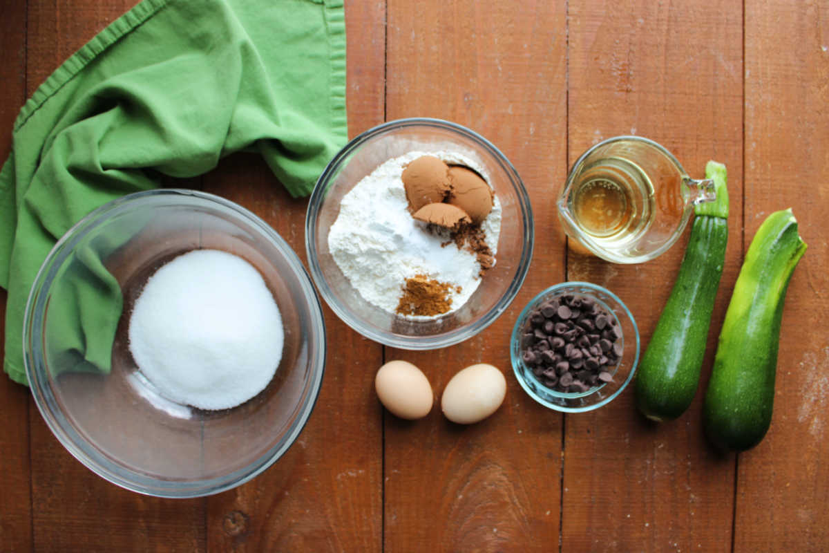 Ingredients including zucchini, sugar, flour, cocoa powder, baking powder, baking soda, salt, cinnamon, oil, eggs, and chocolate chips ready to be made into a chocolate zucchini quick bread. 