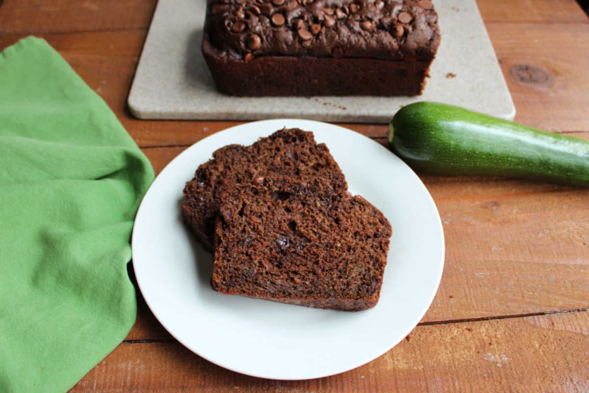 Two slices of chocolate zucchini bread on small plate with a zucchini and a loaf of chocolate zucchini bread in the background.