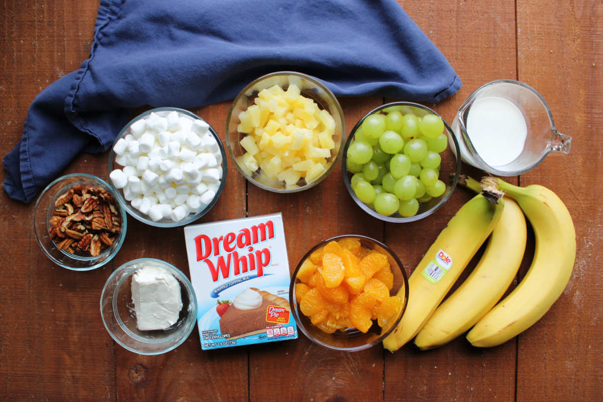 Ingredients including pecans, mini marshmallows, pineapple tidbits, grapes, milk, bananas, mandarin oranges, Dream Whip, and cream cheese ready to be made into grandma's grape supreme salad. 