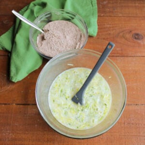 Large glass mixing bowl with wet ingredients including shredded zucchini it in with a smaller bowl of dry ingredients nearby, ready to be incorporated.