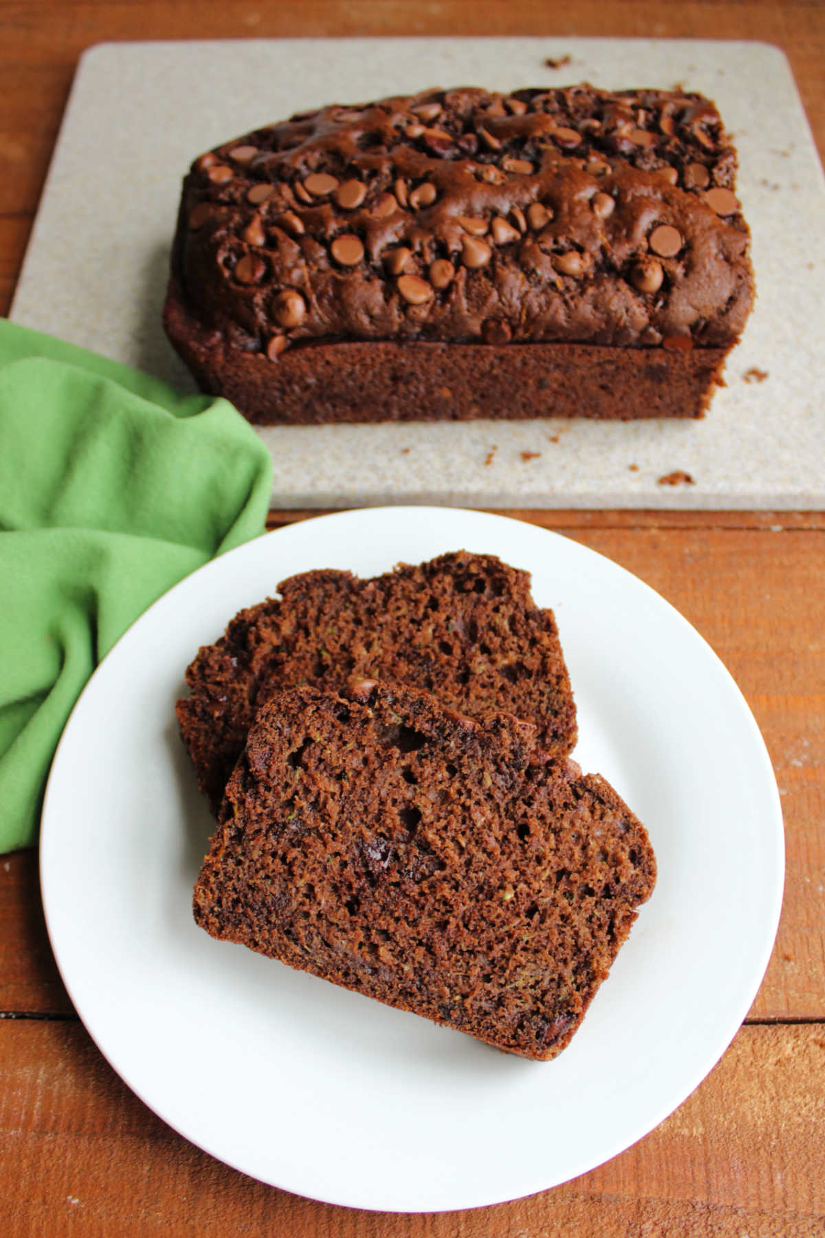 Slices of chocolate zucchini bread with chocolate chips on plate with cutting board holding the remaining loaf of quick bread in the background.