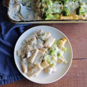 No Boil Chicken Alfredo Bake 9 Dinner plate with a serving of chicken alfredo pasta with broccoli and melted mozzarella cheese on it next to the baking dish it was cooked in.