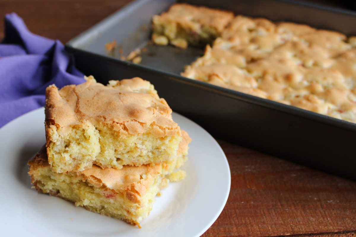 Rhubarb Snack Cake 7 Two squares of rhubarb snack cake stacked on a plate with a pan of the remaining rhubarb snack cake in the background.