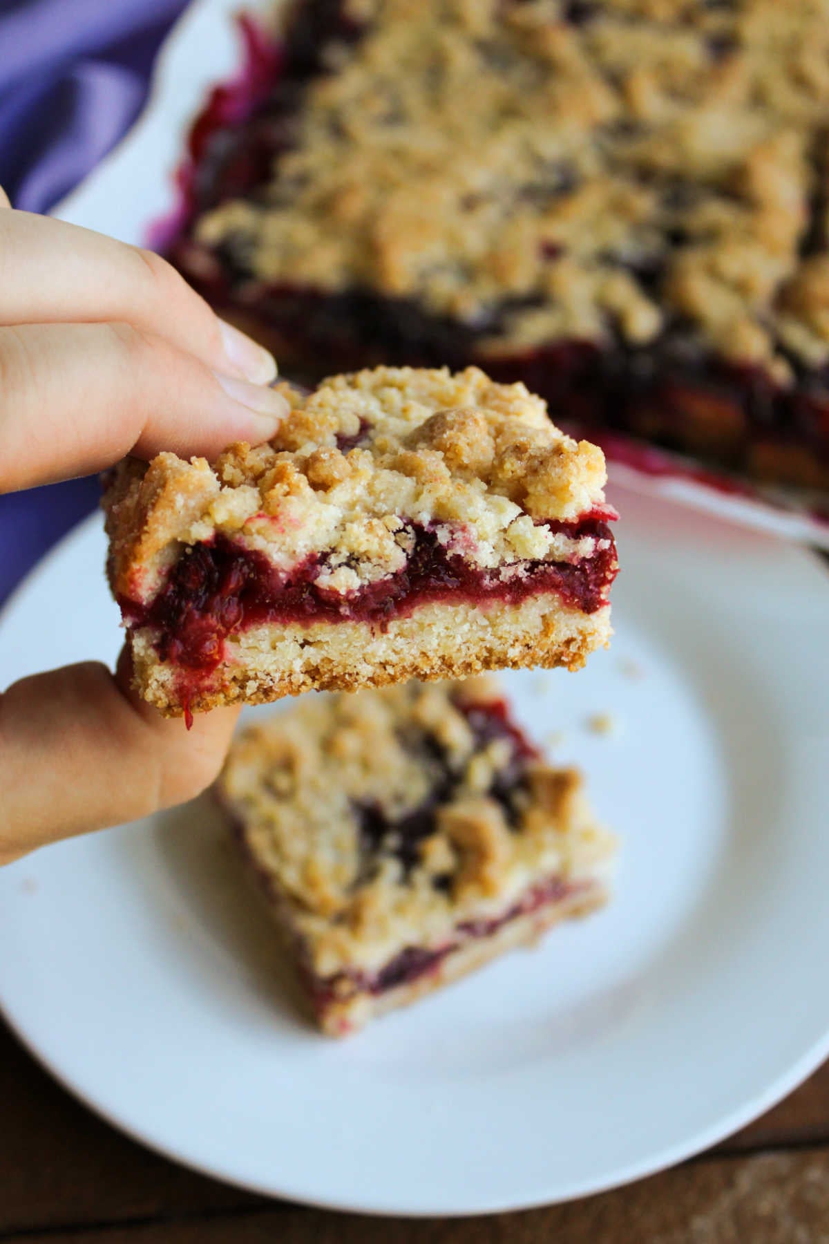 Blackberry Pie Bars 6 Hand holding a blackberry pie bar with a shortbread style crust topped with berry filling and crumb topping, ready to eat.