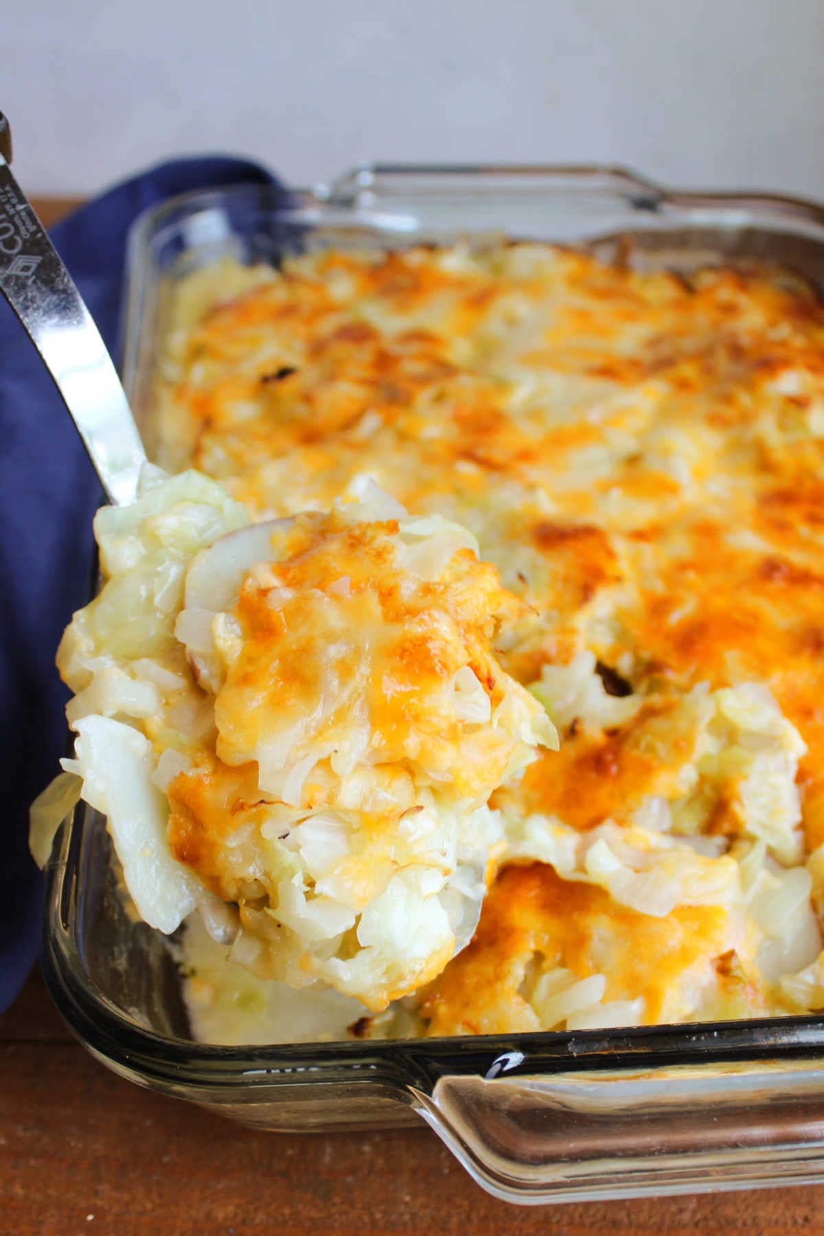 Serving spoon getting a scoop of cabbage and potato casserole out of the baking dish showing the melty cheese, tender veggies, and light sauce. 