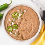 Bowl of creamy homemade refried beans topped with crumbled queso fresco, slices of jalapeno, and some cilantro.