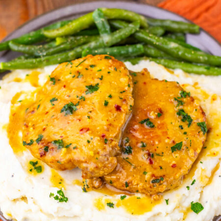 Baked Maple Dijon Pork Chops 1 Close up of a pile of mashed potatoes topped with two maple dijon pork chops with the extra sauce drizzled over the top served with roasted green beans.