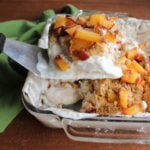 Spatula lifting piece of peaches and cream icebox cake out of pan showing layers of whipped cream, graham cracker crumbs, and diced peaches.