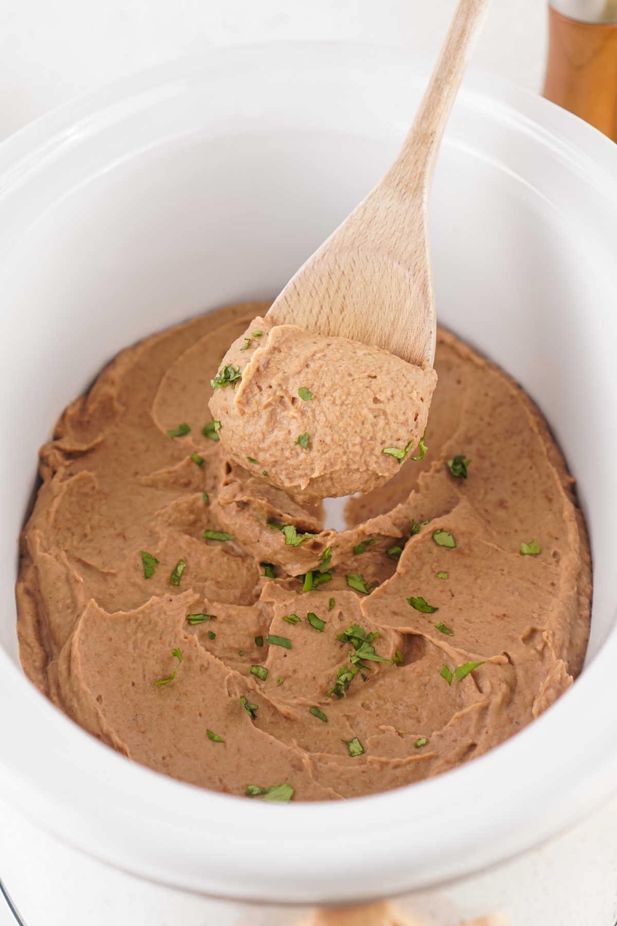 Wooden spoon getting a scoop of homemade refried beans out of the slow cooker. 