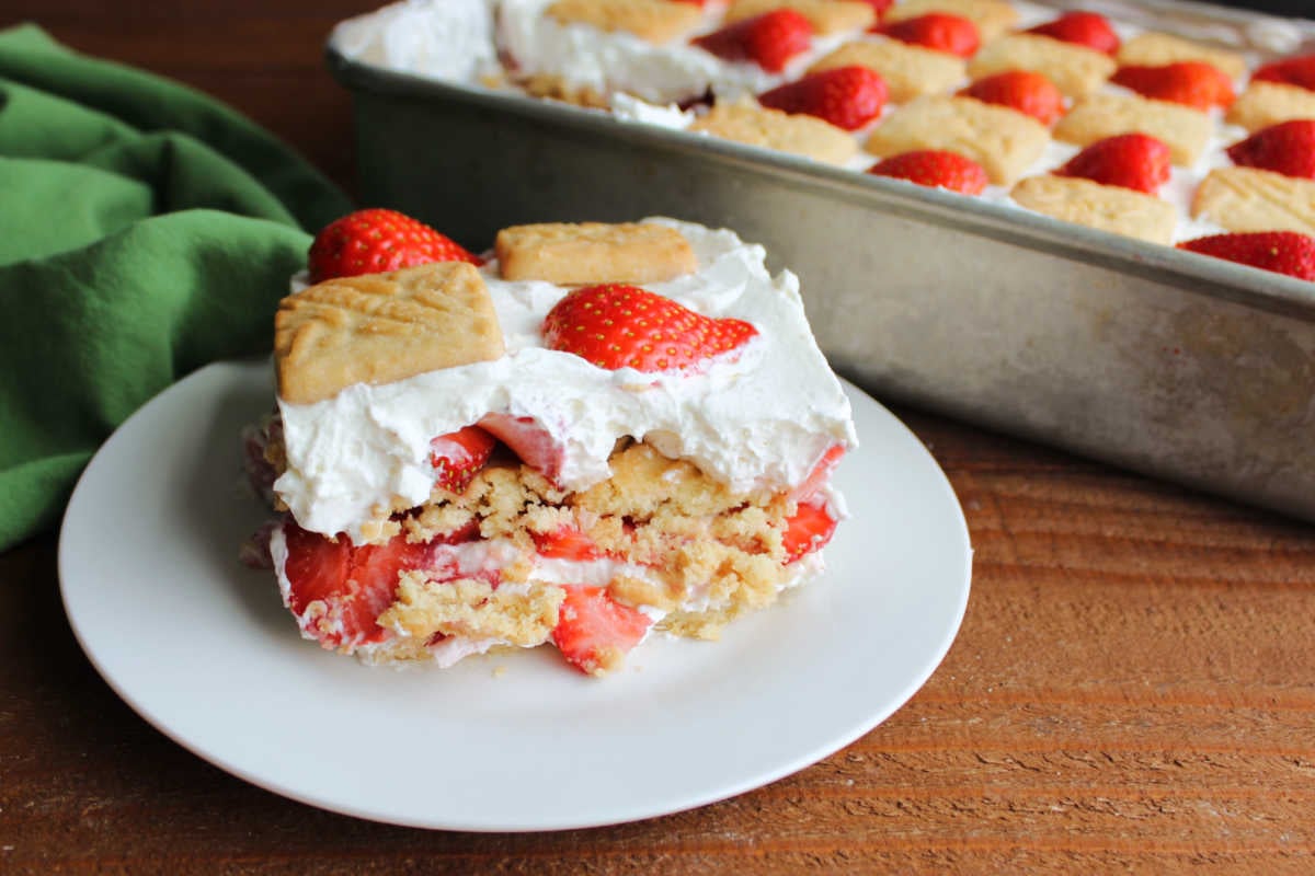Piece of strawberry icebox cake on plate showing layers of cream cheese whipped cream, sliced strawberries, and shortbread cookies. 