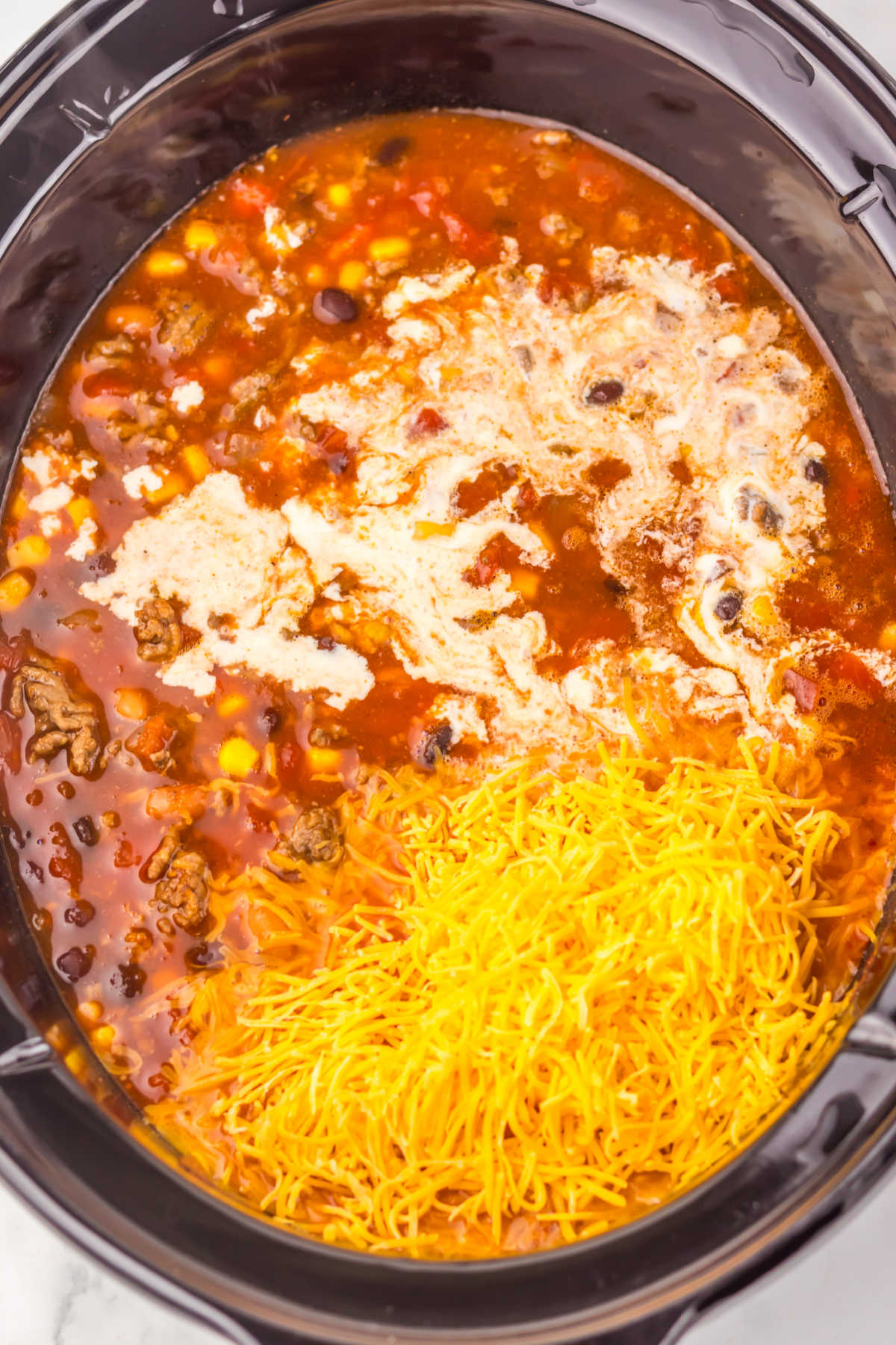 Slow cooer filled with mostly cooked taco soup with some cream and shredded cheese added, ready to be stirred in. 