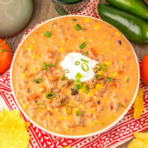Bowl filled with creamy taco soup with ground beef, beans, corn, and tomatoes in cheesy broth topped with a dollop of sour cream and sliced green onions.