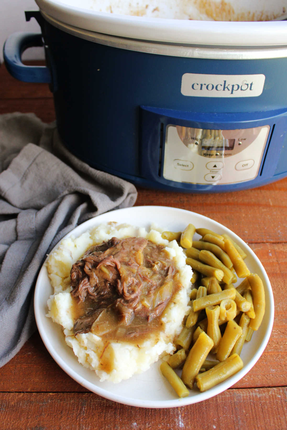 Crockpot filled with Mississippi venison roast next to a dinner plate with some of the meat and gravy served over mashed potatoes with a side of green beans. 