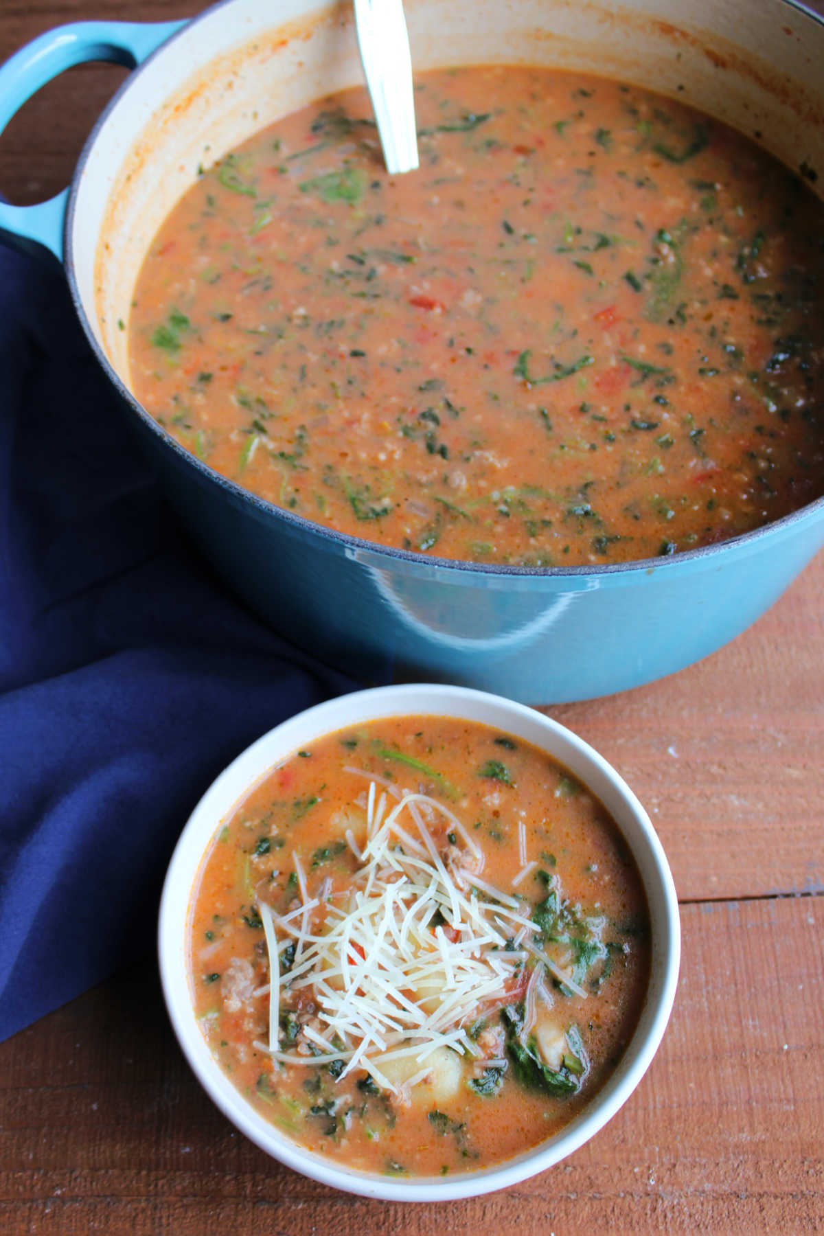 Bowl of Italian sausage soup with spinach and gnocchi topped with cheese next to a large dutch oven with more soup inside. 