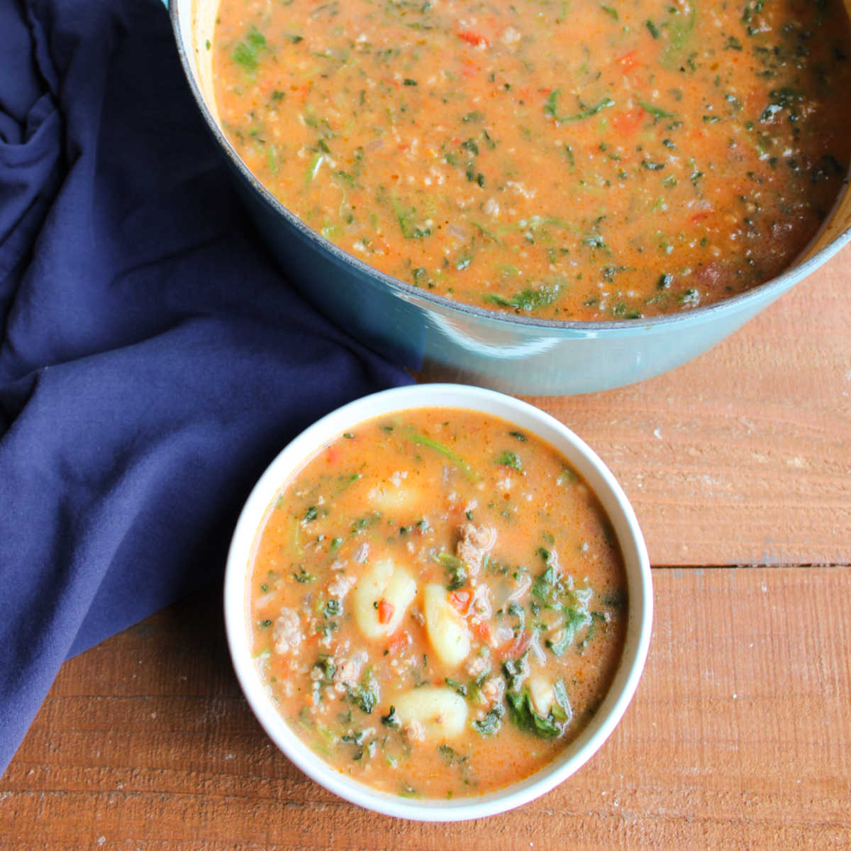 Bowl of creamy tomato soup filled with bits of Italian sausage, gnocchi, and spinach next to soup pot with more inside. 