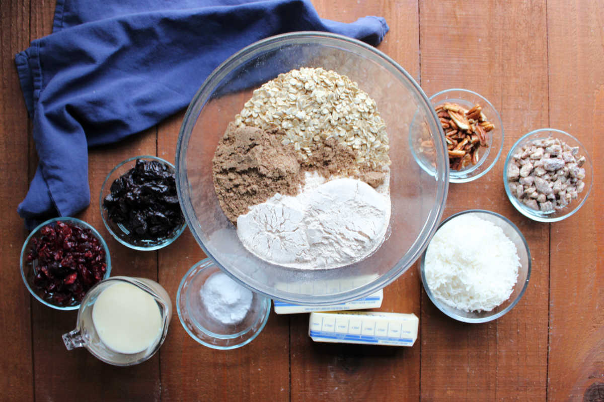 Ingredients including craisins, chopped prunes, ground flax, oats, whole wheat flour, pecans, chopped dates, shredded coconut, butter, baking soda, and sweetened condensed milk ready to be made into trail mix bars. 