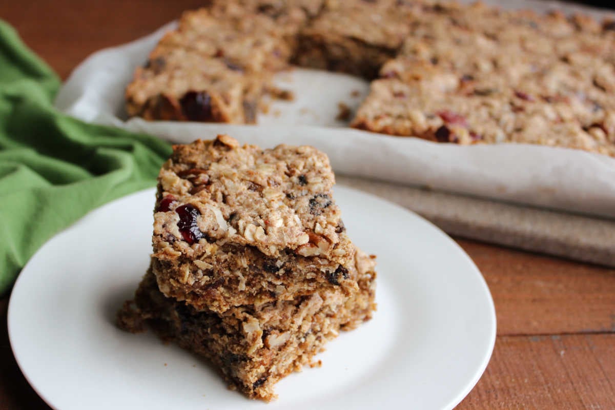 Pieces of chewy trail mix bars with oats, dried fruit, and nuts in front of cutting board with remaining bars in the background. 
