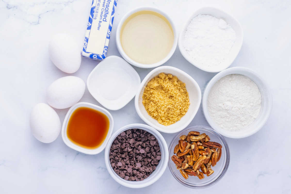 Ingredients including butter, flour, powdered sugar, corn syrup, pecans, brown sugar, mini chocolate chips and eggs ready to be made into chocolate pecan pie bars. 