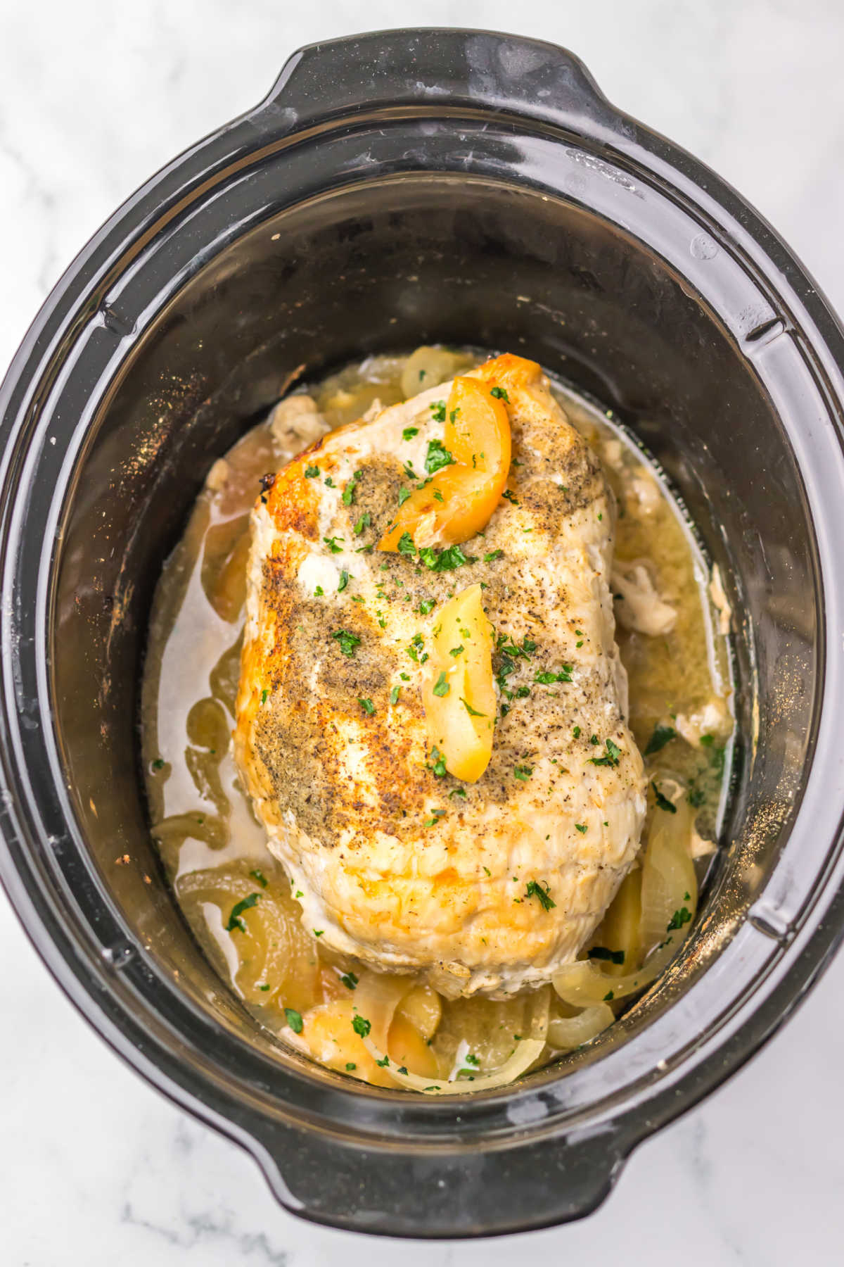 Crockpot with cooked boneless turkey breast inside showing tender cooked meat, cooking juices, and softened apples and onions. 