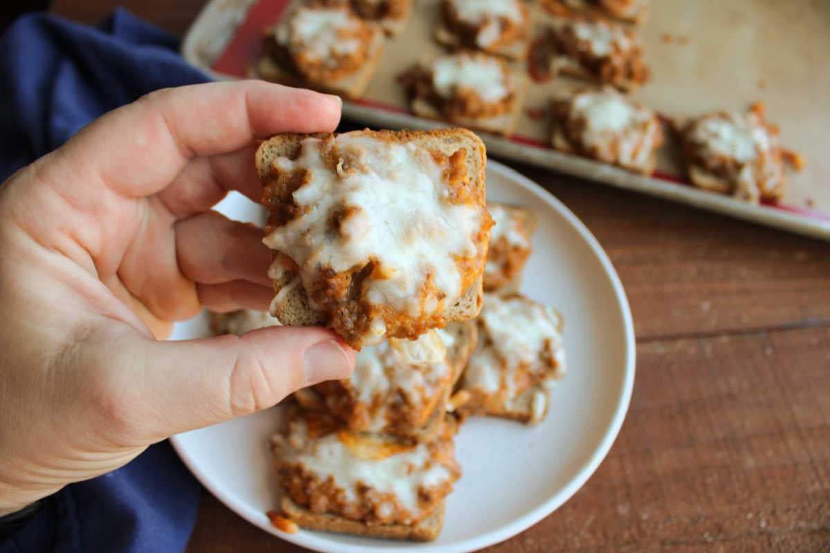 Jeff's Pizza Bites 7 Hand holding a cocktail rye topped with sausage, pizza sauce and cheese mixture with melted mozzarella cheese on top, ready to eat.