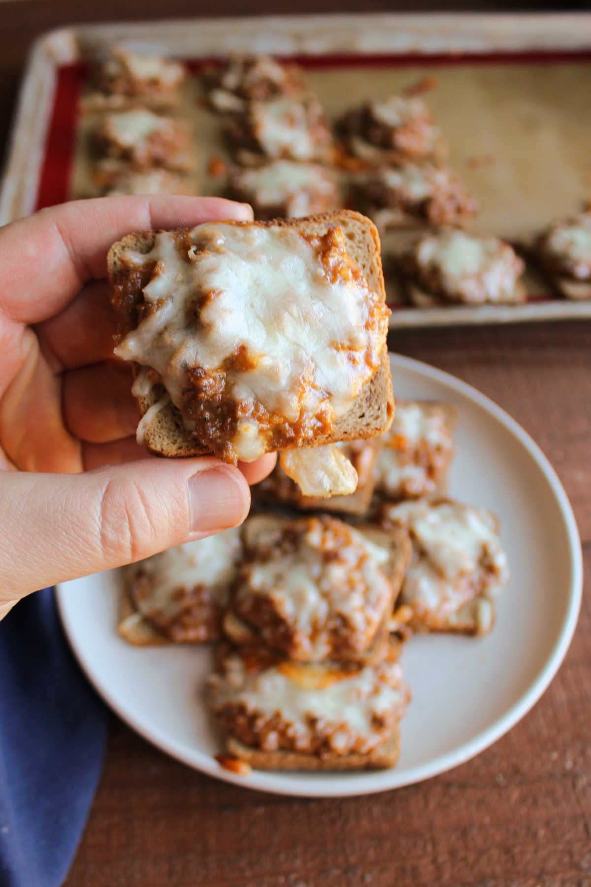 Jeff's Pizza Bites 9 Hand holding a pizza snack with cheesy sausage and pizza sauce mixture on a square of cocktail rye bread, ready to eat.