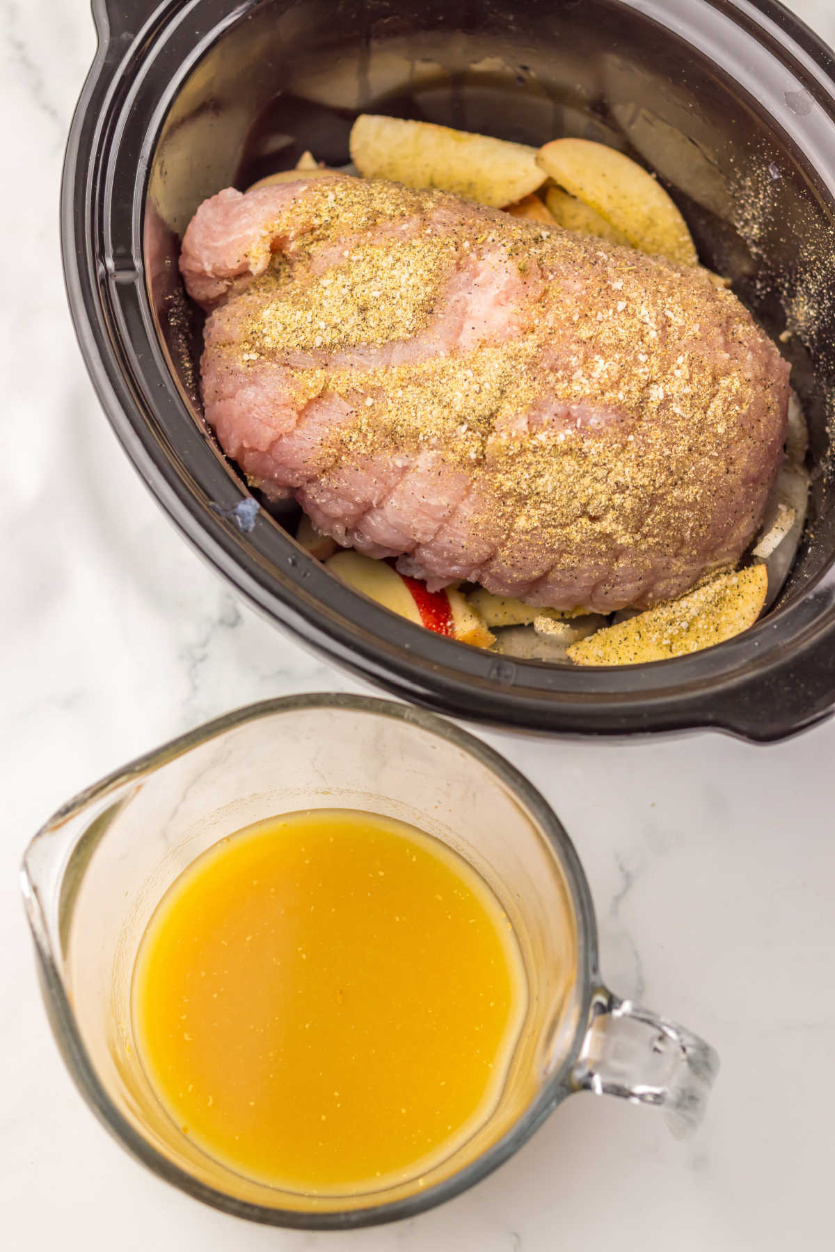 Seasoned boneless turkey breast sitting on top of apple and onion slices in a slow cooker with a large measuring cup that contains the gravy mixture nearby. 