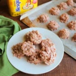Plate of no bake strawberry cookies with oats next to a cookie tray with more cookies setting up on wax paper and the container of strawberry milk powder.