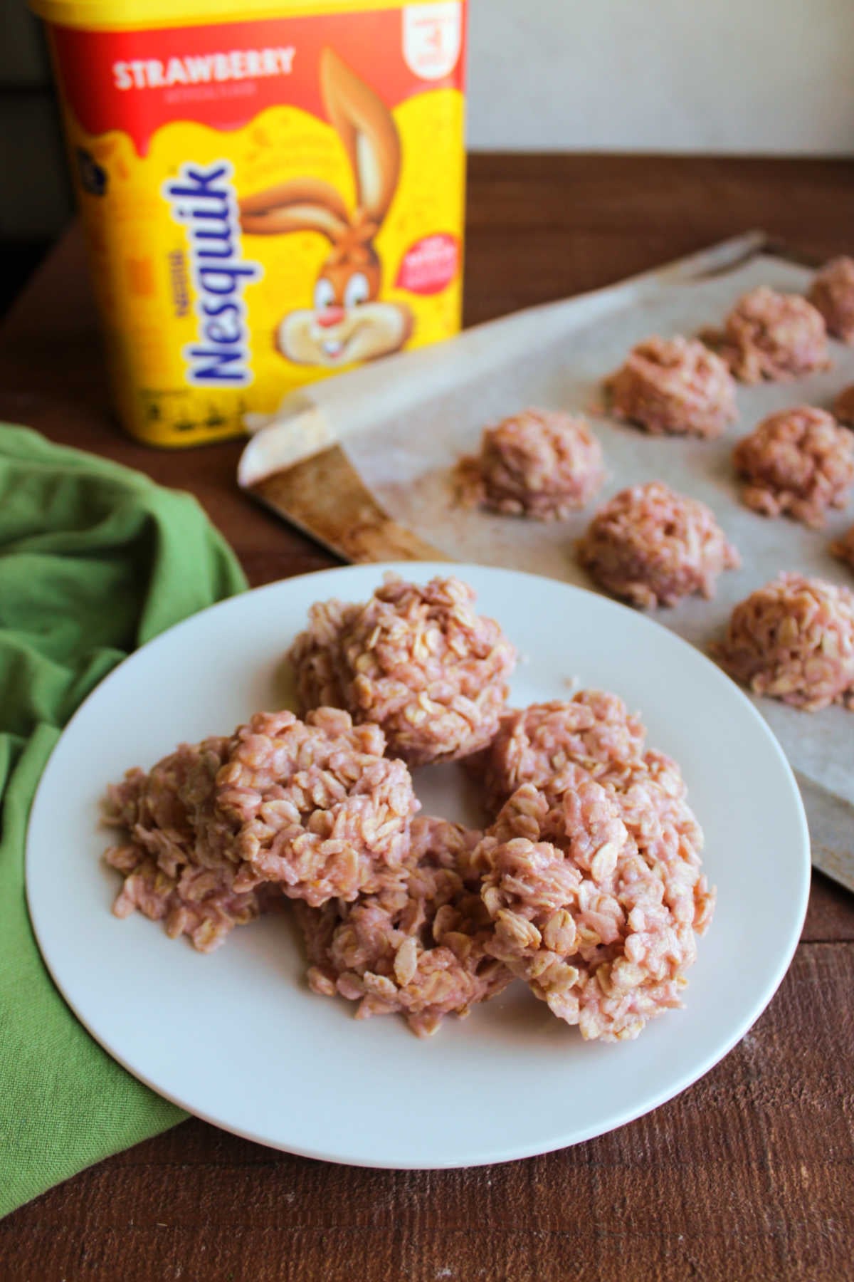 Plate of no bake strawberry cookies next to a tray of more cookies and a container of strawberry milk powder. 