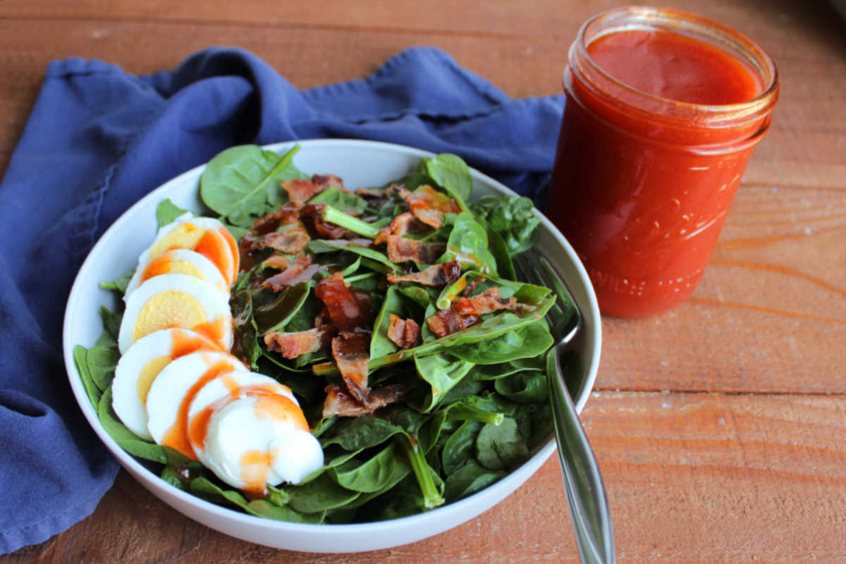 Bowl of spinach salad with bacon and hard boiled egg next to a jar of homemade catalina salad dressing. 